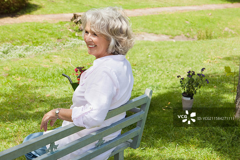 一位老年女性坐在花园的长椅上，旁边有一辆装满红花的手推车和几盆紫色植物。图片素材