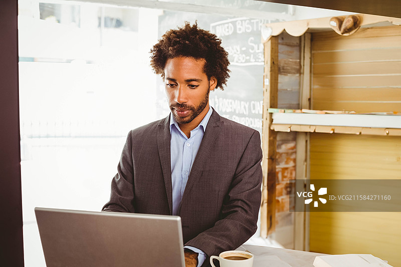 一位穿着深色西装的非裔美国男性在咖啡店里用笔记本电脑打字，旁边有一杯咖啡。图片素材