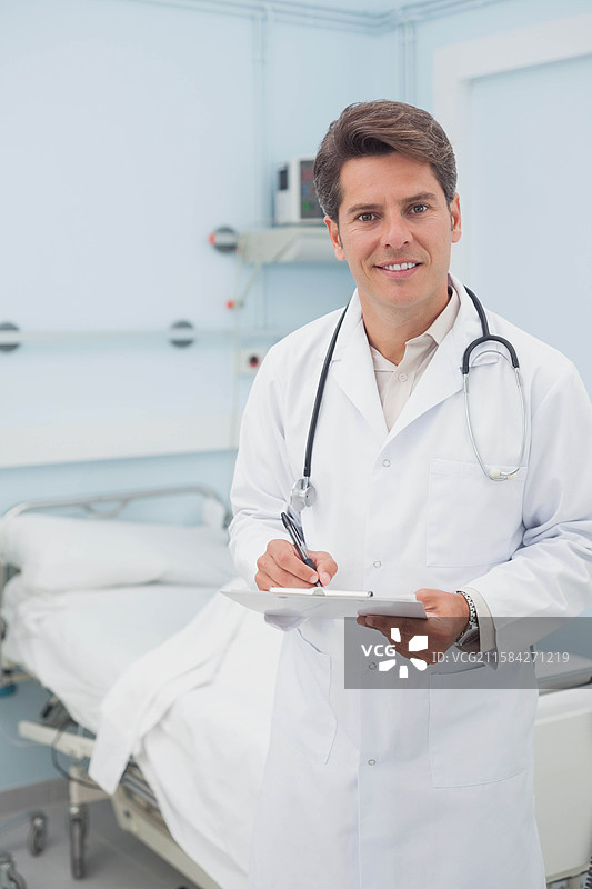 微笑的男医生在病房的病床旁用笔在夹板上写字，身旁有听诊器。图片素材