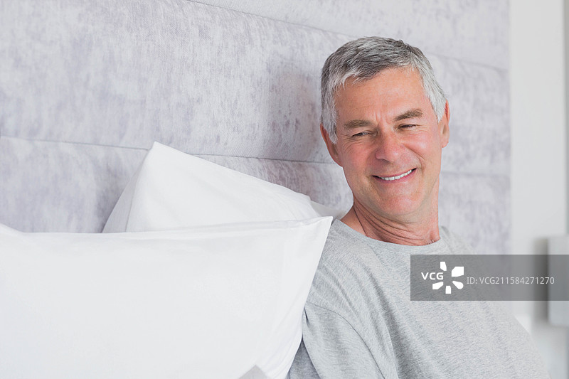 穿着灰色T恤的老年男子坐在床上，白色的枕头和床头板，留白空间图片素材