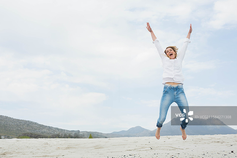 穿着扣子衬衫和卷起裤脚的牛仔裤的女人在沙地上跳跃，天空中有空白空间。图片素材