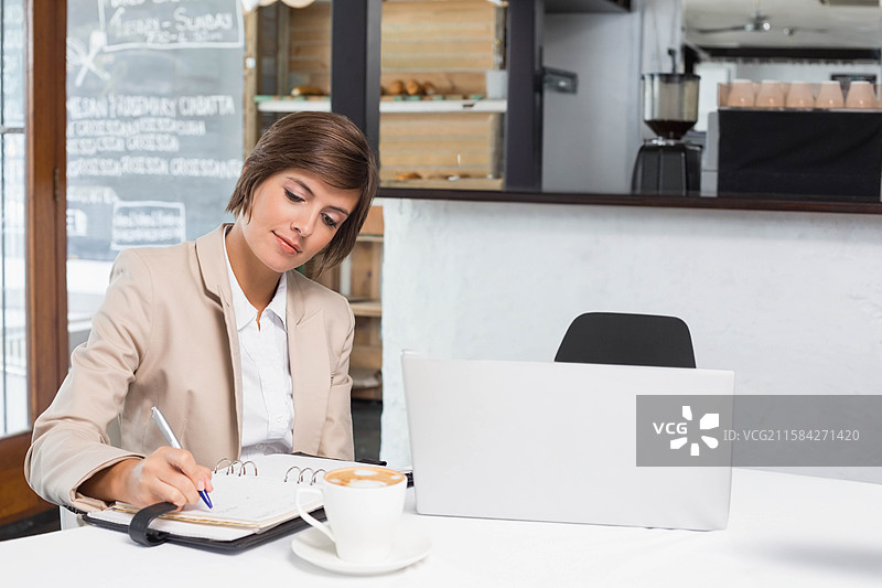 穿着西装的女商人在咖啡馆的桌子上写笔记，旁边有笔记本电脑和咖啡杯，留有空白空间图片素材