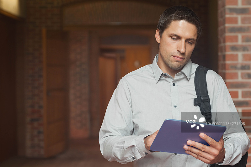 穿着衬衫、背着背包的男子在砖砌走廊中使用平板电脑，留有空白空间。图片素材