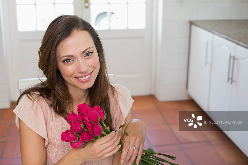 女人在明亮的家用厨房中，胸前抱着一束鲜艳的粉色玫瑰花，靠近玻璃门。图片素材