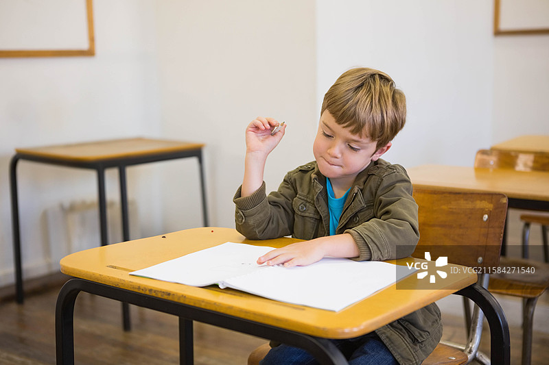 在教室里，穿着夹克的学龄男孩坐在桌子旁，专注于手中的练习册，握着铅笔。图片素材