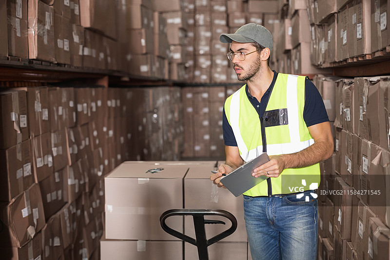 男性仓库工人手持夹板和铅笔，在仓库过道检查箱子，留白空间图片素材