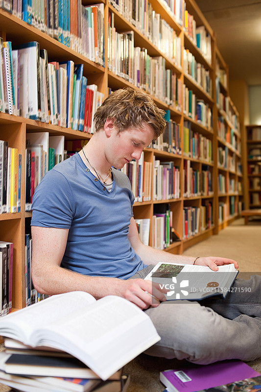 男子坐在图书馆地板上，阅读教科书，旁边是木制书架和紫色笔记本。图片素材