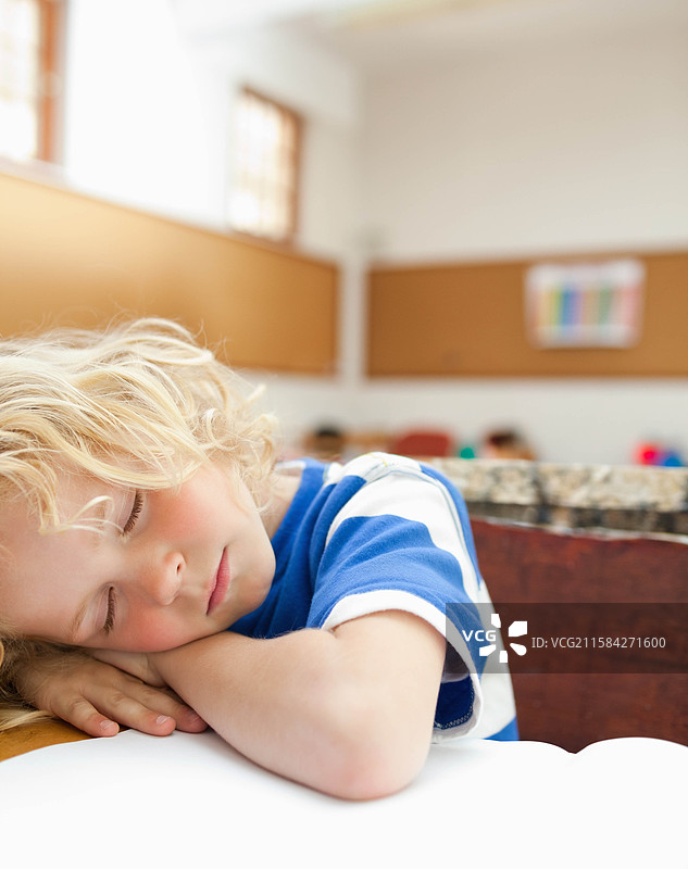 学校年龄的男孩把头靠在折叠的手臂上，趴在教室的木桌上，桌上有一张空白的笔记本纸。图片素材