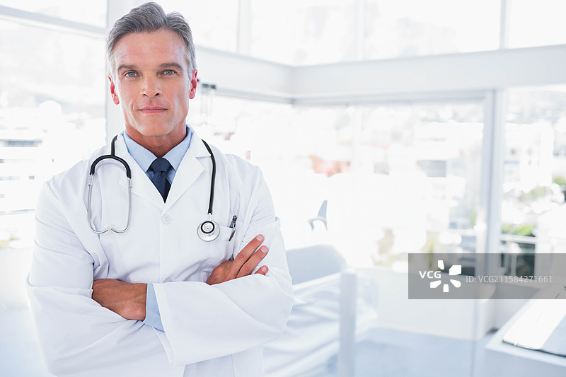 成熟男性医生在医疗诊所中双臂交叉，佩戴听诊器，穿着实验室外套。图片素材