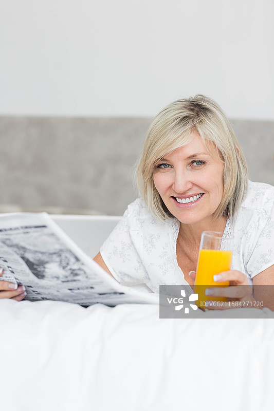 女人在卧室的床上靠着，手里拿着一杯橙汁，正在阅读报纸，留有空白空间。图片素材