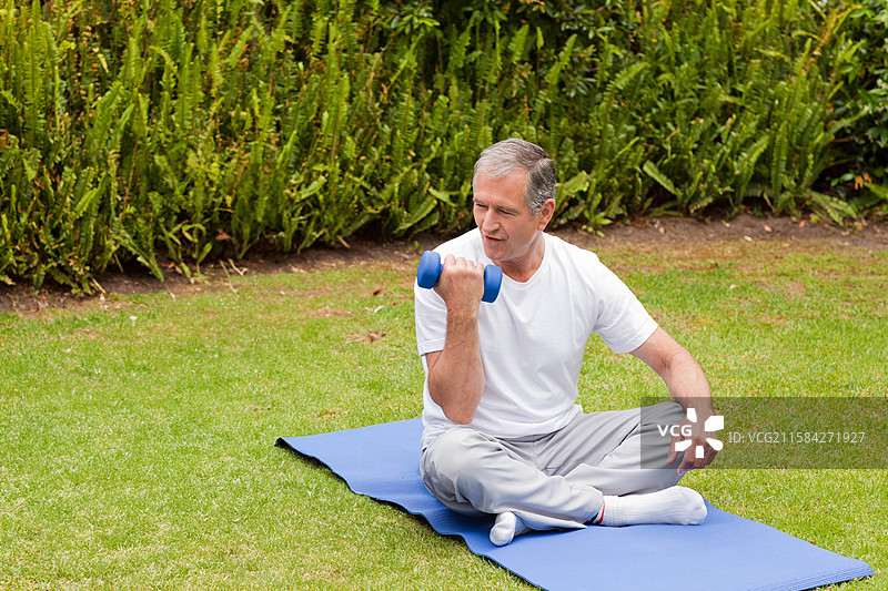 老年男子在草坪上的蓝色运动垫上用蓝色哑铃做二头肌弯举图片素材