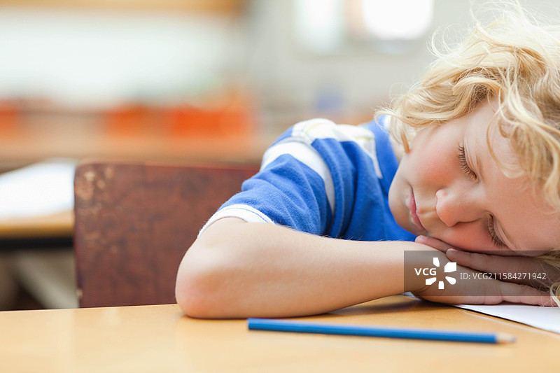 男孩在教室的木桌上休息，头靠在桌子上，手臂靠近蓝色铅笔和纸张。图片素材