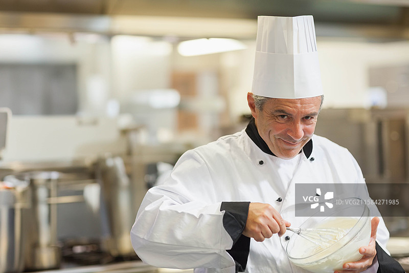 男厨师戴着高帽，用金属打 whisk 在碗中搅拌混合物，餐厅厨房，留白空间图片素材