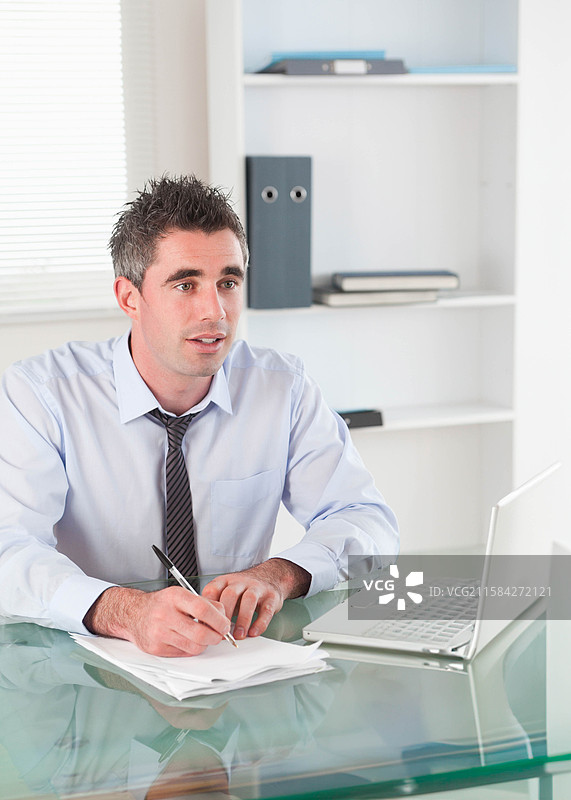 身穿商务服装的男子在玻璃桌上写字，旁边有打开的笔记本电脑和文件夹。图片素材