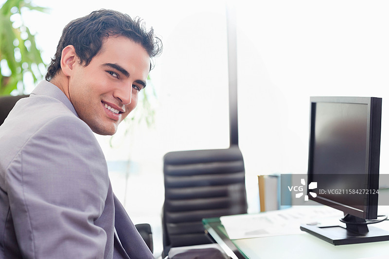 男人微笑着转动办公室玻璃桌旁的椅子，桌上有显示器、书籍和文件，留有空白空间。图片素材