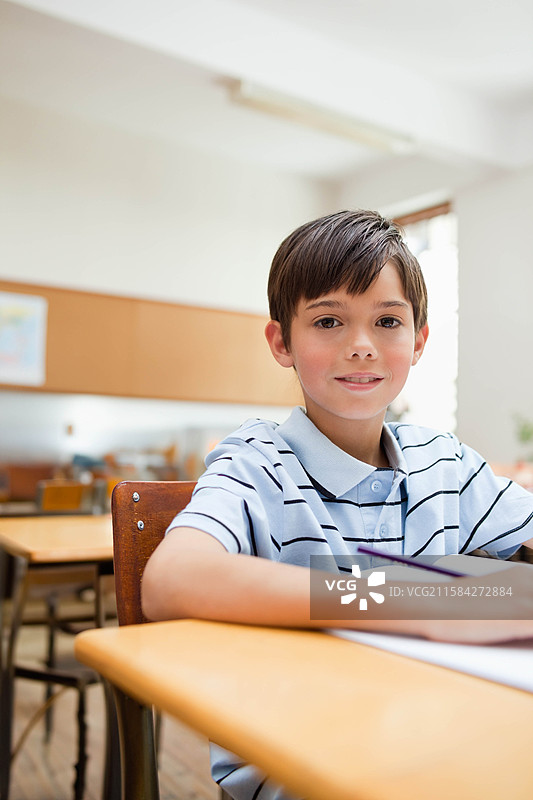 白人男学生坐在教室的木桌前，用铅笔在打开的笔记本上写字图片素材