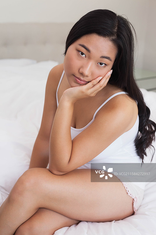 穿着粉色吊带衫的韩国女性盘腿坐在床上，床单为白色，旁边有一个玻璃床头柜。图片素材