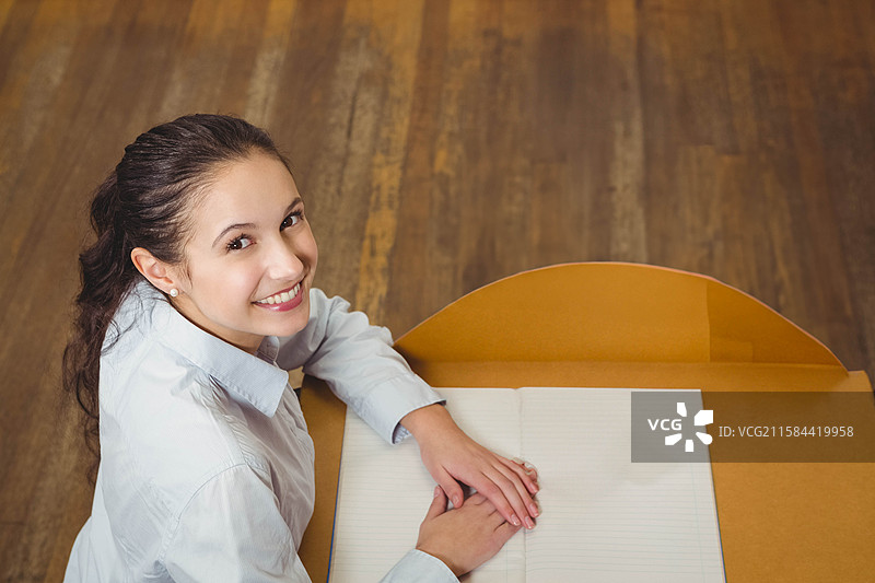 穿着白色衬衫的女性坐在木桌前，面带微笑地看着镜头，旁边有一本打开的笔记本，留有空白空间。图片素材