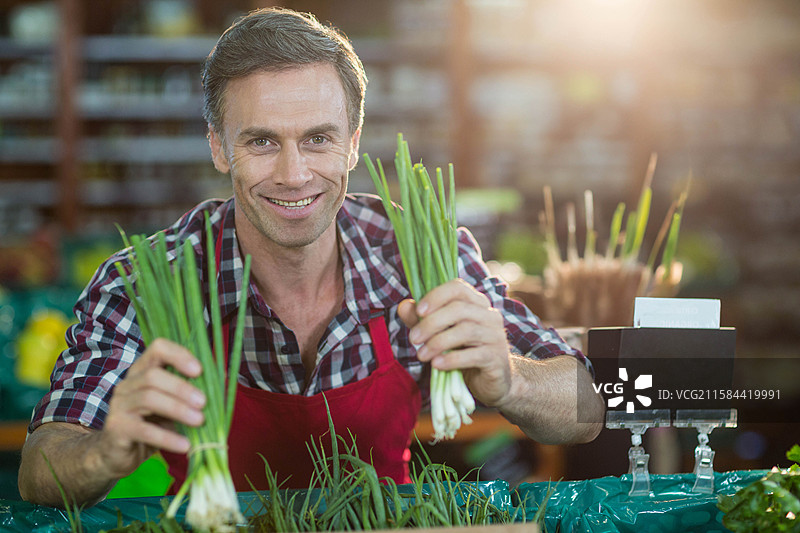 成熟的男摊贩在超市的塑料覆盖桌子后微笑，手里拿着青葱图片素材