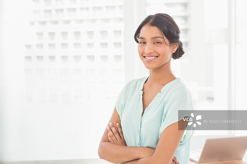 穿着浅蓝色 blouse 的亚洲专业人士在办公室里微笑着站着，旁边有一台笔记本电脑。图片素材