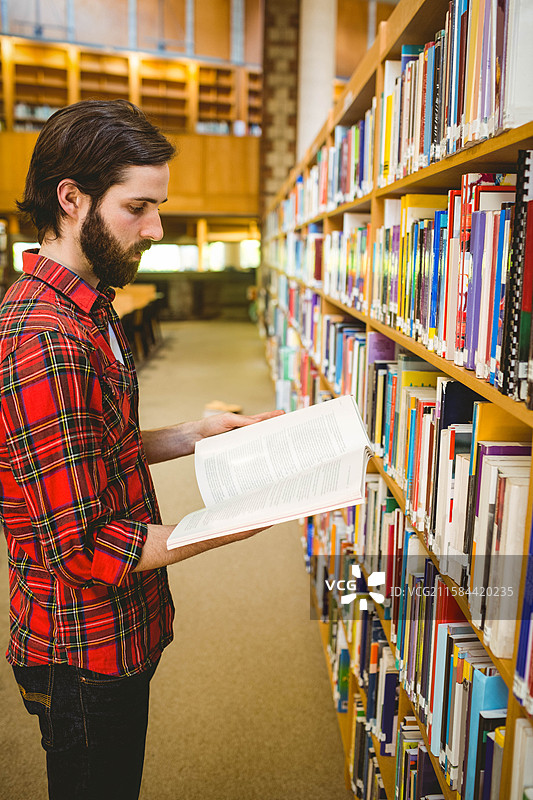 一名成年男性站在图书馆的木质书架之间，手中拿着一本打开的书，旁边留有空白空间。图片素材