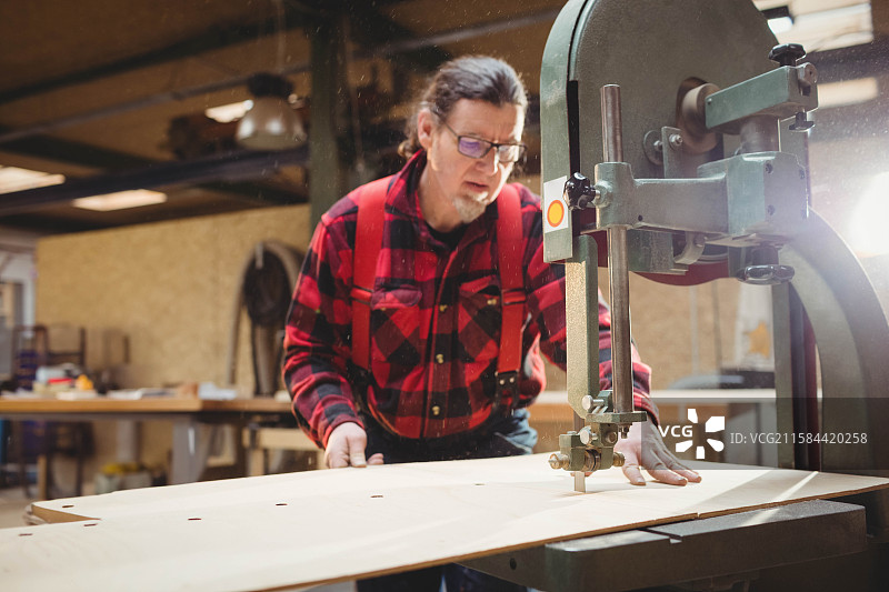 成熟男性穿着法兰绒衬衫，在木工车间中引导胶合板通过立式带锯，周围满是锯屑。图片素材