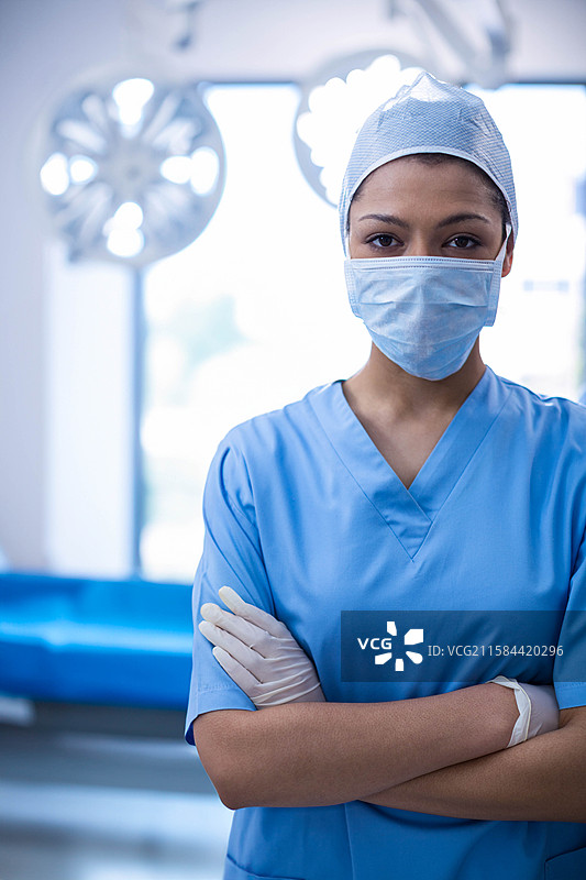 穿着手术服的非裔美国女性外科医生，双臂交叉，站在手术室的灯光下。图片素材