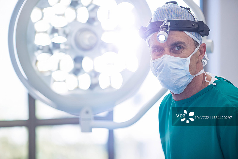 中年外科医生站在LED灯下，穿着绿色手术服和头灯，在手术室内。图片素材
