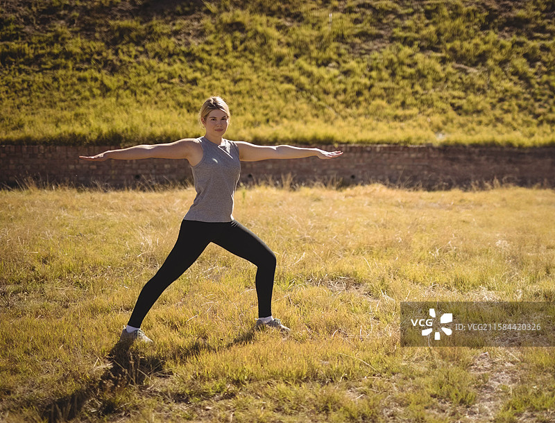 女人在阳光下的砖墙旁，干燥的黄色草地上做着战士二式瑜伽姿势。图片素材