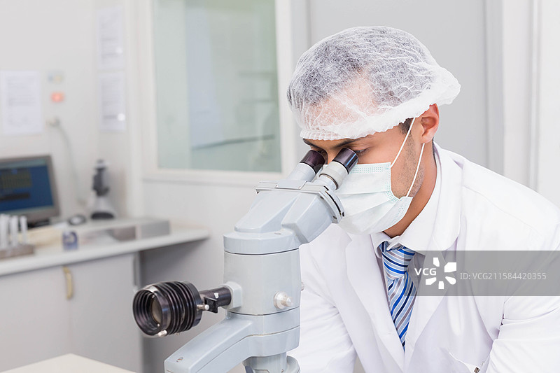 实验室技术人员在工作台上通过光学显微镜观察，旁边有电脑显示器和移液器架。图片素材