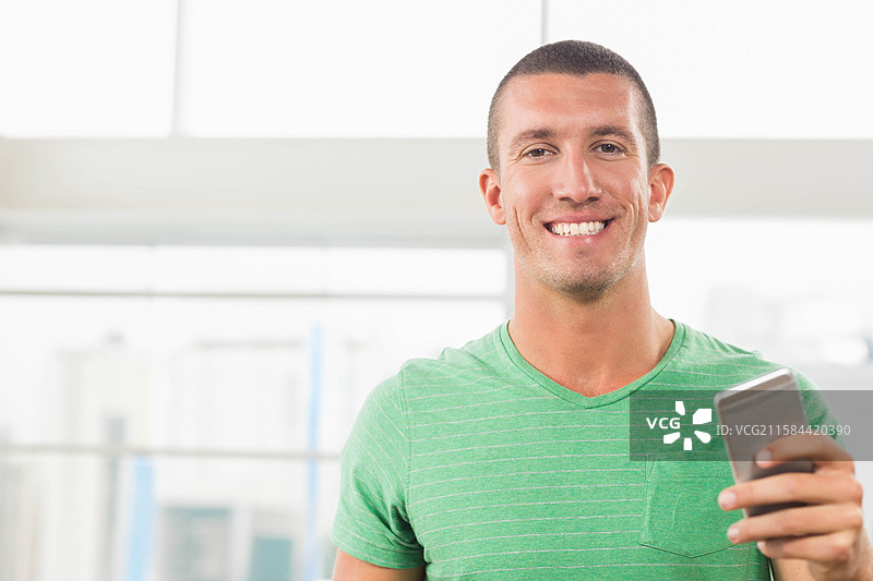 一位成年男性微笑着将智能手机举至胸前，站在现代化的房间里，背景是白色墙壁和玻璃栏杆。图片素材