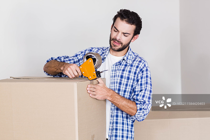男子在空房间里使用胶带分配器封闭一个大纸箱，身后有一个装满物品的纸箱。图片素材