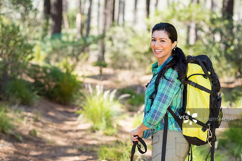 穿着衬衫、背着黄色背包的西班牙裔女性在森林小径上徒步旅行，手持登山杖，留有空白空间。图片素材