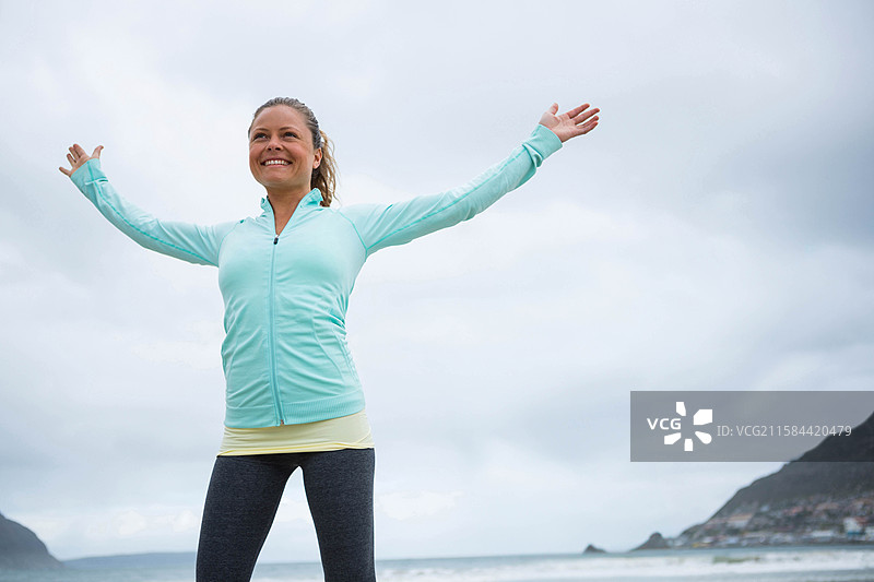穿着运动服的女性在海边沙滩上伸展双臂，天空多云。图片素材
