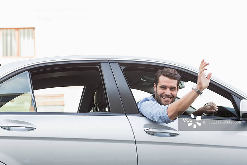 男性从银色轿车的驾驶窗外伸出手，挥手致意，手腕上戴着手表，街道上有空白空间。图片素材