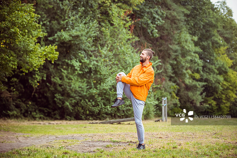 穿风衣的胡子男人在公园里做膝盖到胸部的拉伸，旁边有健身器材，留有空白空间图片素材