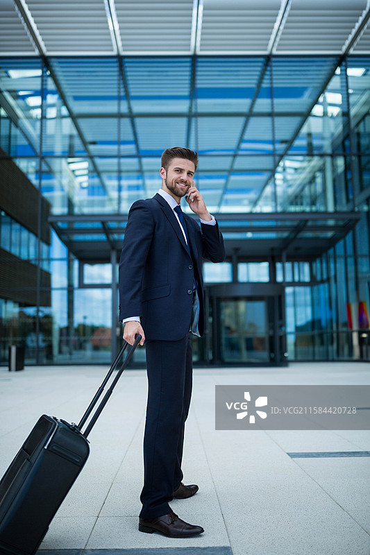 穿着得体的男士在办公广场上用智能手机通话，同时拉着行李箱，周围有玻璃顶棚。图片素材