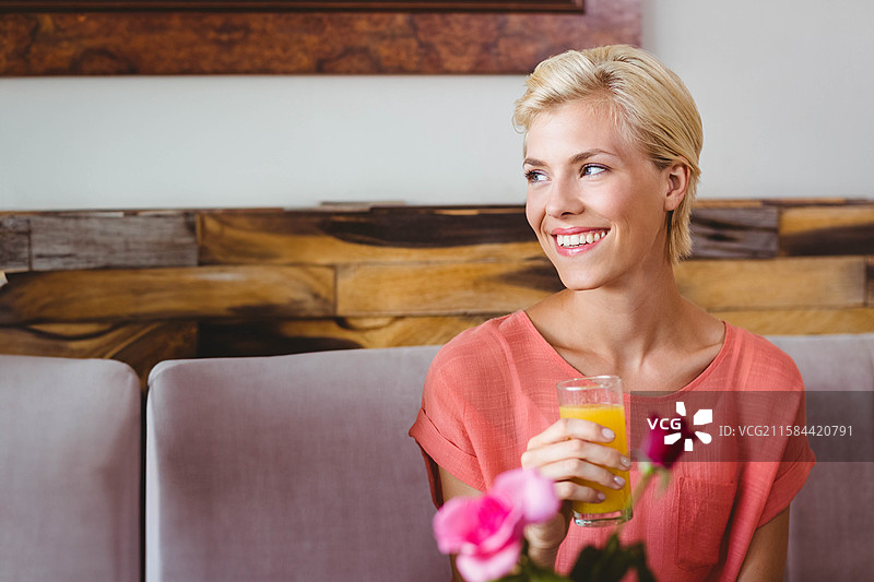 女性微笑着坐在咖啡馆休息区的长椅上，手里拿着一杯橙汁，周围有鲜花。图片素材
