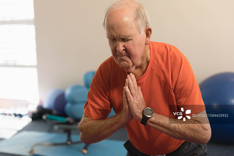 穿着橙色衬衫的老年男子在健身房中双手合十，身旁有蓝色健身球。图片素材