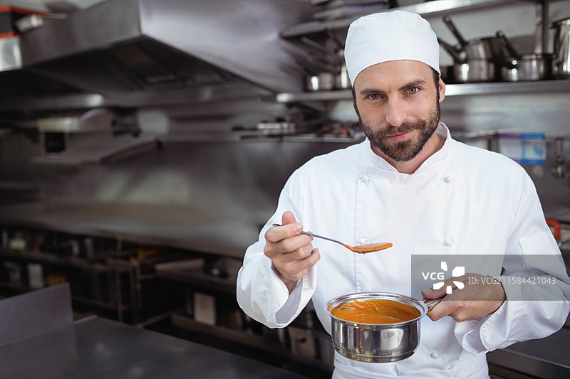 专业男性厨师在商业厨房中拿着平底锅并品尝番茄汤，留有空白空间图片素材
