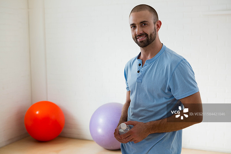 男子在健身工作室里手持水瓶，旁边是红色和薰衣草色的稳定球，留有空白空间。图片素材