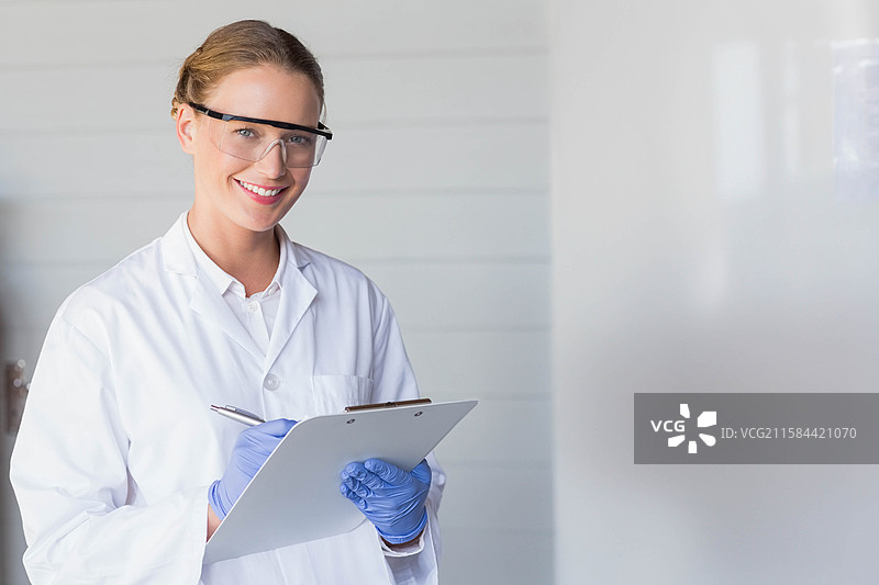 穿着实验室外套、护目镜和手套的女性技术人员在实验室中使用夹板和笔，留有空白空间。图片素材