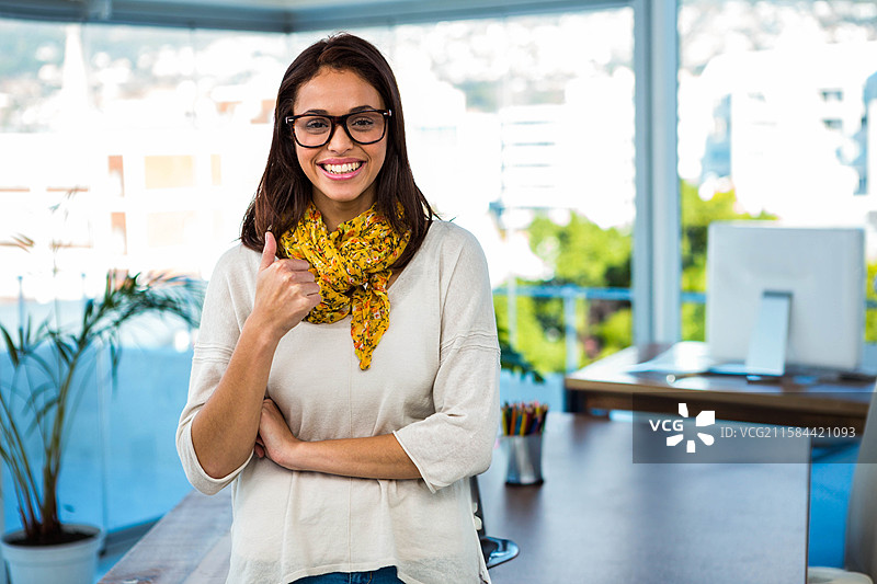 女性在办公室空间中站立，竖起大拇指，旁边是带显示器的桌子和大窗户。图片素材