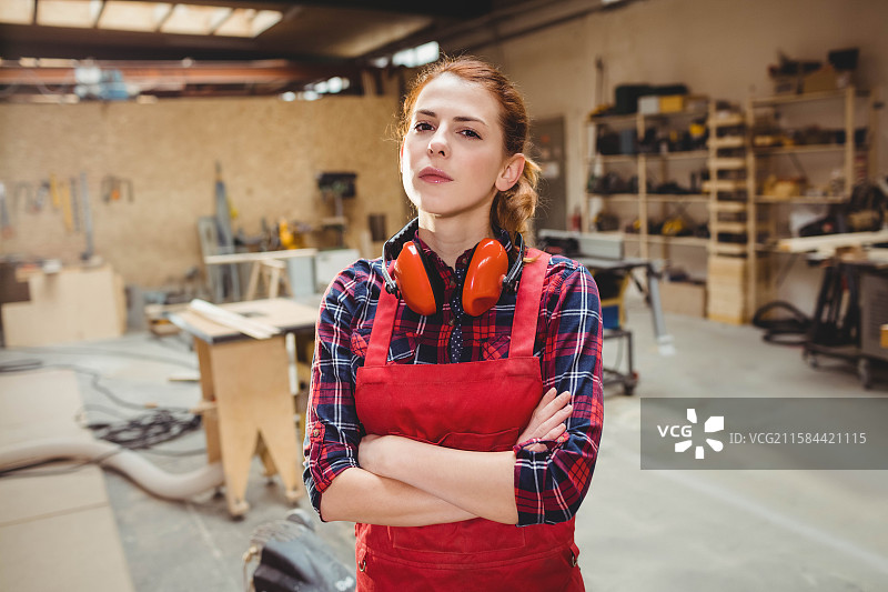 穿着红色围裙和耳罩的女性木工，站在工作台和木材之间的工作坊里图片素材