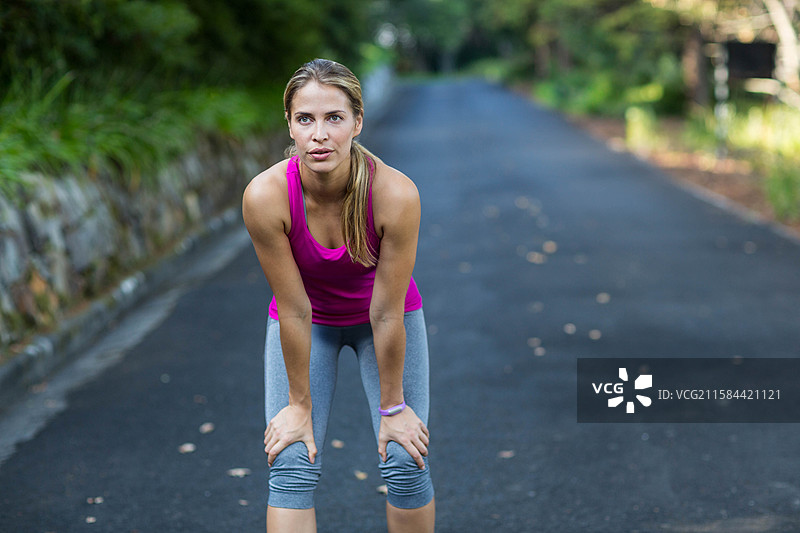 穿着粉色背心和健身追踪器的女性跑者在树木成荫的道路上喘息。图片素材