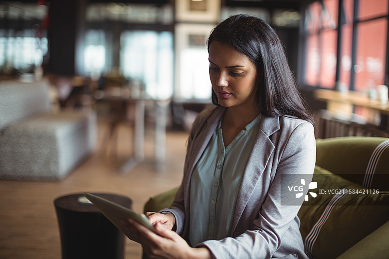 穿着商务服装的亚洲女性坐在橄榄绿色椅子上，手持平板电脑，位于咖啡馆休息区。图片素材