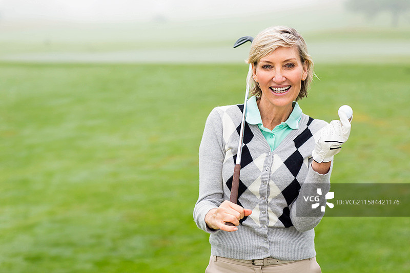 一位年长女性高尔夫球手将球杆搭在肩上，手套握着球，站在球道上，留有空白空间。图片素材