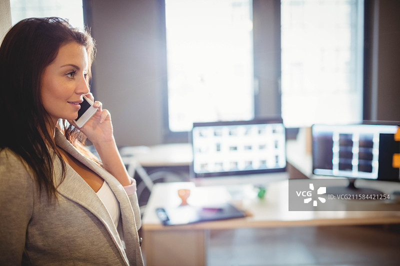 穿着西装的女性站在办公桌旁，手持智能手机靠在耳边，留有空白空间图片素材