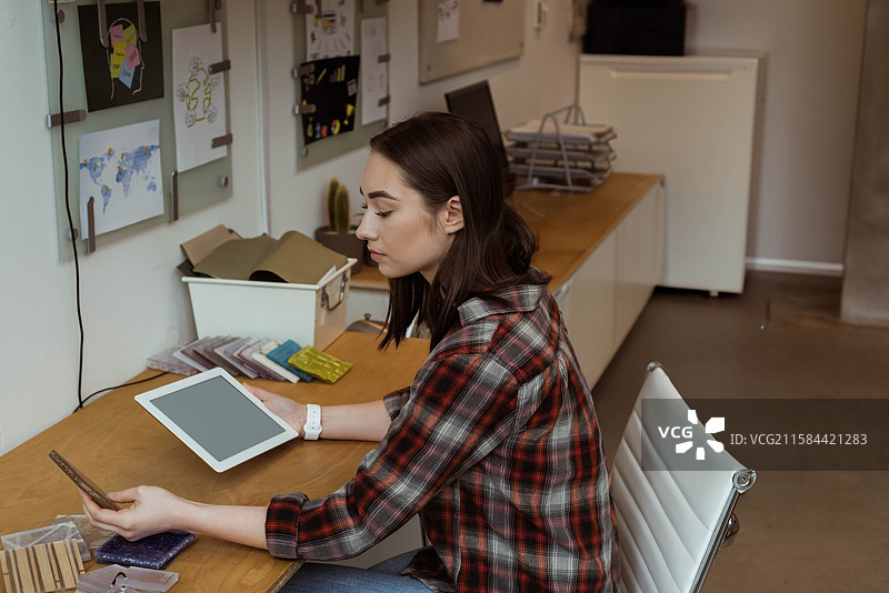 二十多岁的女性坐在木桌前，手持平板电脑并使用智能手机，在设计工作室内。图片素材