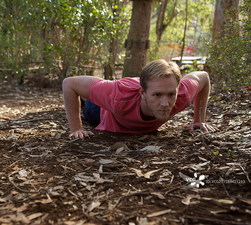 穿着红色衬衫的成年男性在森林地面上做俯卧撑，周围有干枯的树叶和树枝。图片素材